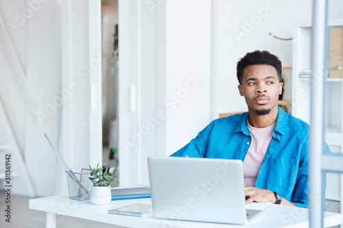 African young businessman in blue casual shirt sitting at his workplace with laptop with pensive look