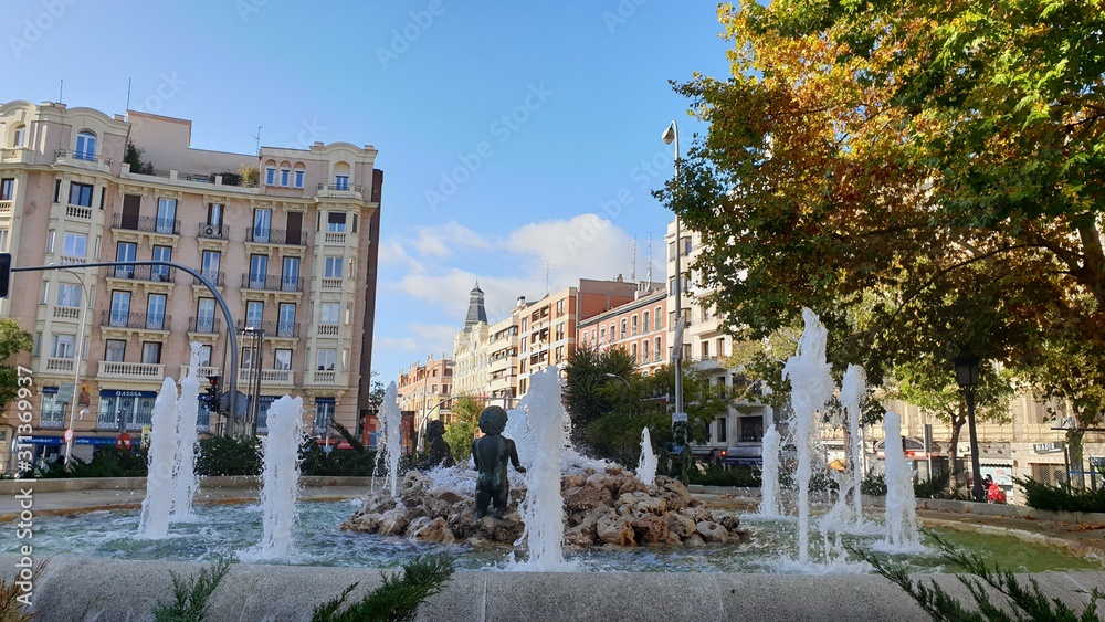 Madrid Autumn Square: a fountain against a background of yellow-green trees, high-rise buildings and a blue sky with white clouds, in daylight Spain Madrid