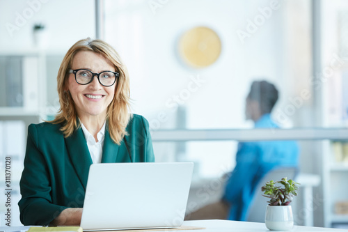 Portrait of successful mature businesswoman in eyeglasses sitting at the table in front of laptop and smiling at camera at office