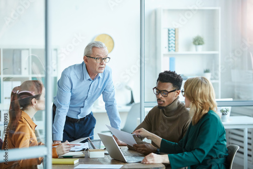 Mature businessman in eyeglasses standing and explaining the business strategy to young business people at the table at meeting at office