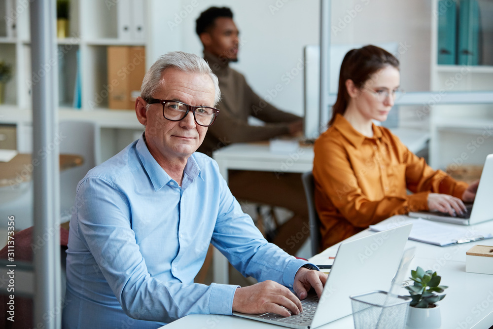 Portrait of mature businessman in eyeglasses typing on laptop computer while sitting at desk with his colleagues working in the background at office Portrait of mature businessman in eyeglasses typing on laptop computer while sitting at desk with his colleagues working in the background at office