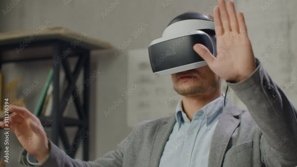 Modern man engineer-designer in the office with large windows stands in a virtual reality helmet uses gestures to manage the project without leaving the office. Construction control. Design project Modern man engineer-designer in the office with large windows stands in a virtual reality helmet uses gestures to manage the project without leaving the office. Construction control. Design project