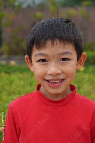 The lovely Asian boy in red is smiling at the park with green background.