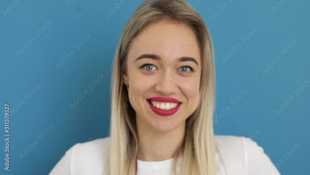 Big smile blonde beautiful young woman looking to the camera and winking. Blue background. White t-shirt. Close up. Video footage Big smile blonde beautiful young woman looking to the camera and winking. Blue background. White t-shirt. Close up. Video footage