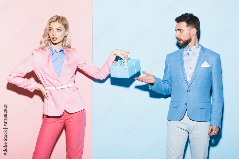 attractive woman holding gift and handsome man pointing with hand on pink and blue background attractive woman holding gift and handsome man pointing with hand on pink and blue background