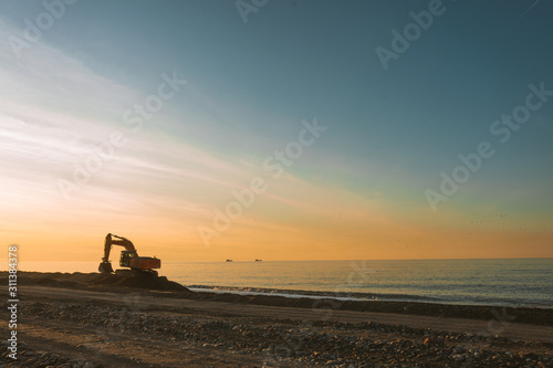 Excavator loads the excavation onto a truck  hydraulic are heavy construction equipment consisting of an arrow a bucket and a cabin on a rotating platform.On the beach with the sea and the setting sun