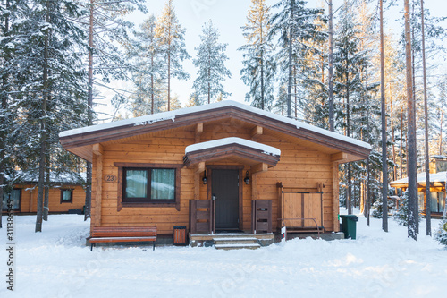 One-storey house made of wooden beams among the trees of the winter forest. Treetops lit by low sun