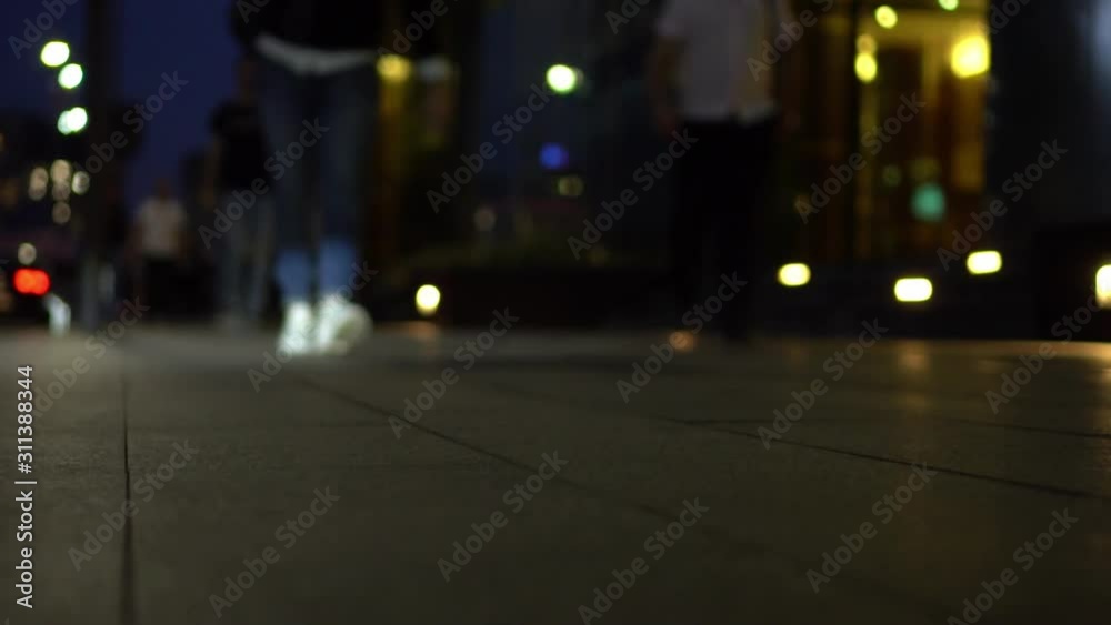 Defocused view of a city street in the evening. A man in blue jeans and white sneakers walks along the sidewalk. People walking behind him in the background. City lights. Night life. Slow Motion Defocused view of a city street in the evening. A man in blue jeans and white sneakers walks along the sidewalk. People walking behind him in the background. City lights. Night life. Slow Motion