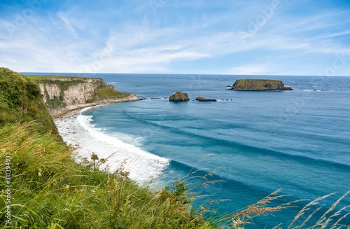 Northern Irish coastline in Carrick-a-rede in Ballycastle. scenic landscape of grass hillsides, rock cliffs and still waves from the Irish sea extending the coastline.