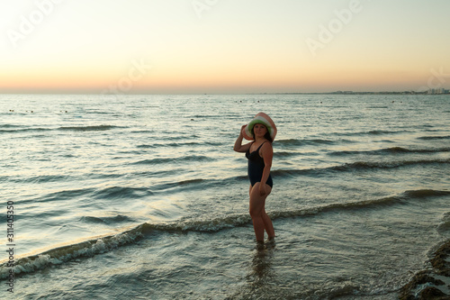 A woman in a hat walks along the seashore at sunset.