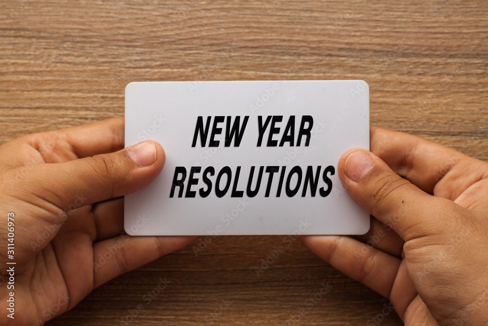 New Year Resolutions written on card with hand holding it with wood background New Year Resolutions written on card with hand holding it with wood background
