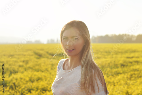Portrait of young woman with long hair in strong afternoon backlight sun, blurred field with yellow flowers background