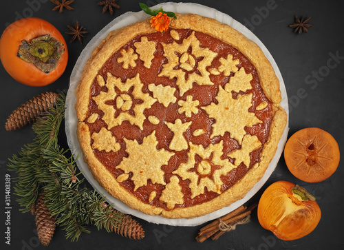 Delicious homemade persimmon jam pie. The cake is decorated with Christmas figures from cookies. Next to the pie are persimmon slices and a Christmas tree. Top view.