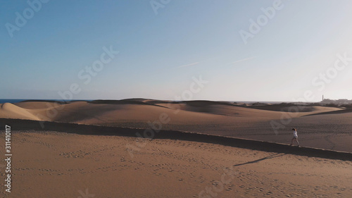 Aerial view is a side view of the dune along which a beautiful young woman walks. The golden hour at sunset or sunrise in the desert. Maspalomas, Gran Canaria