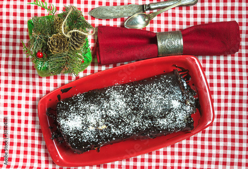 Traditional french chrismtas long cake or yule log, cutlery, red napkin with ring and gift box on the checkered tablecloth. Flat lay