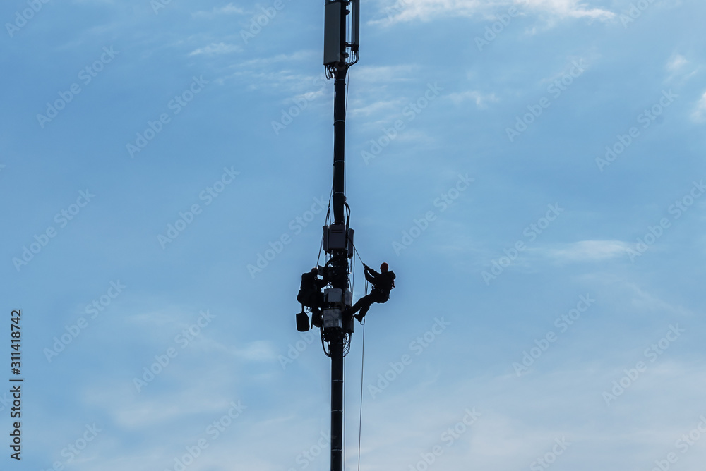 Installers service equipment on a communications pole high above the ground. Backlit silhouettes