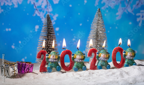 the numbers for the year 2020 stand in the snow with a pine branch decoration and a little imp sit on the left site with a christmas tree in the back on a wooden texture background with shining stars