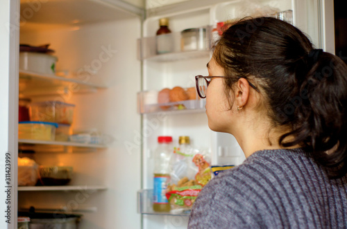 A teenage girl in a gray sweater and glasses with her hair in a bun stands at the open refrigerator and examines the products. The little girl is thinking what she can eat. Selective focus. Blurred ba