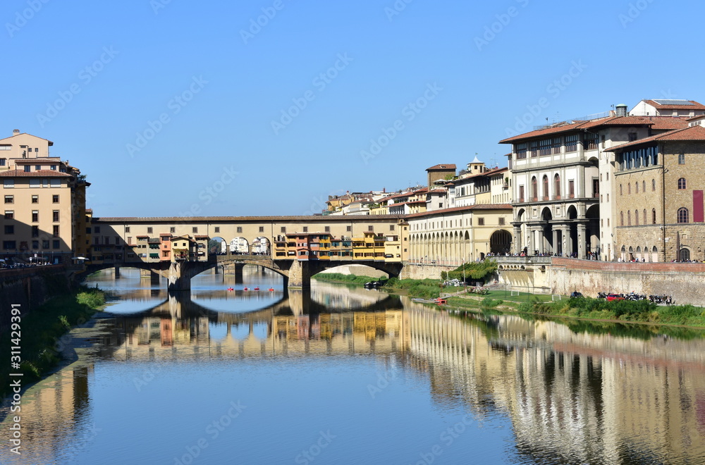View of Ponte Vecchio with blue sky and water reflections from bridge over the Arno River. Florence, Italy.