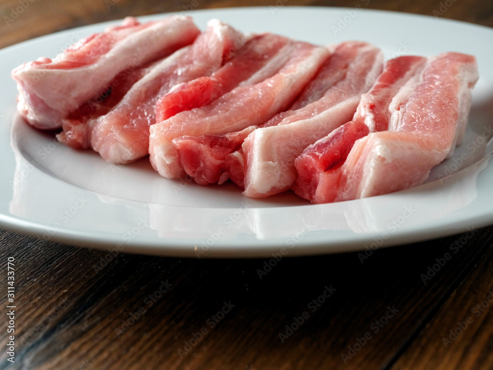 Fresh uncooked pork belly slices on a white plate. Wooden table surface. Fresh uncooked pork belly slices on a white plate. Wooden table surface.