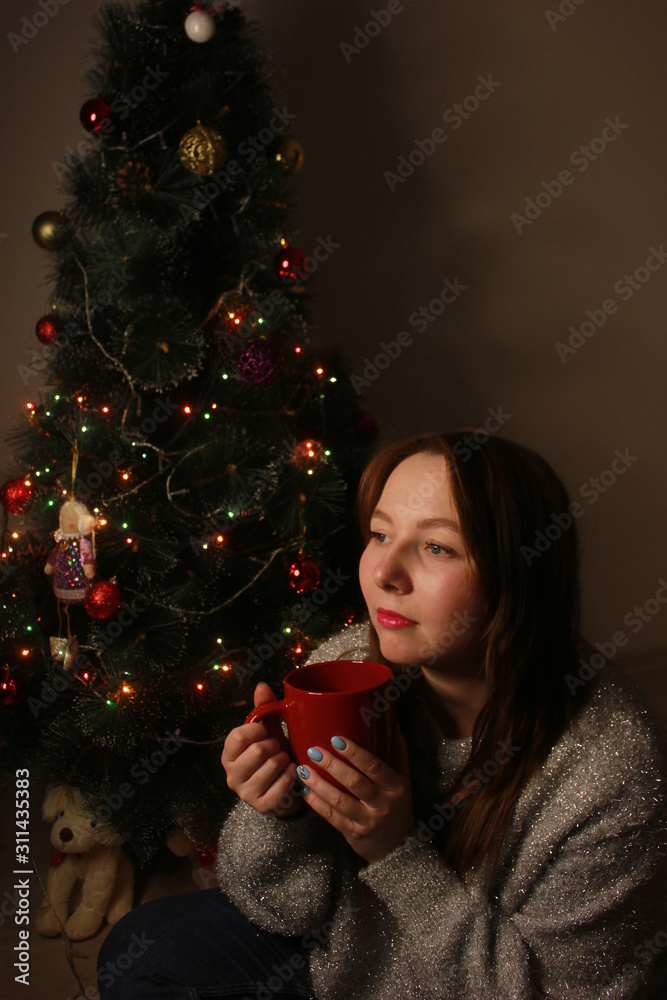 Beautiful young woman drinking tea at the Christmas tree. Beautiful girl celebrates Christmas with a cup of cocoa. Beautiful young woman drinking tea at the Christmas tree. Beautiful girl celebrates Christmas with a cup of cocoa.