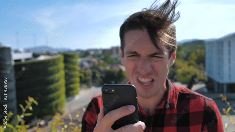 Man is angry with the message that he read in the smartphone. Aggressive Young Man in plaid shirt Using Smartphone outdoor on urban city background. 