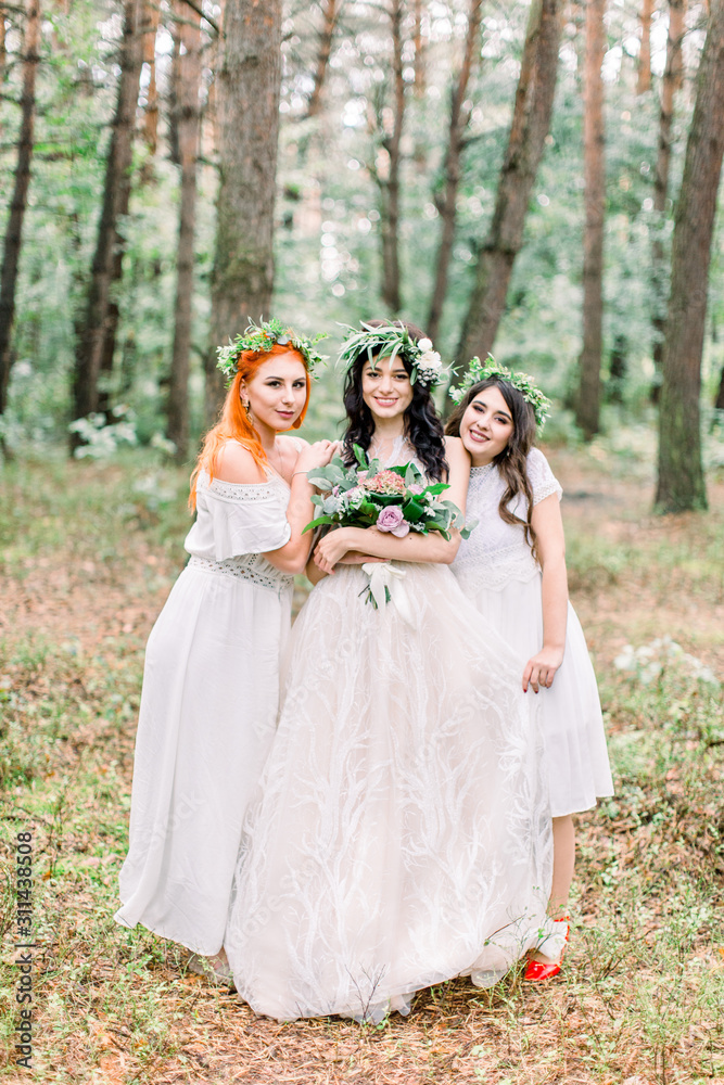 Rustic wedding outdoors. The bride in a white dress and floral wreath standing with her two bridesmaids in green forest. Rustic wedding outdoors. The bride in a white dress and floral wreath standing with her two bridesmaids in green forest.