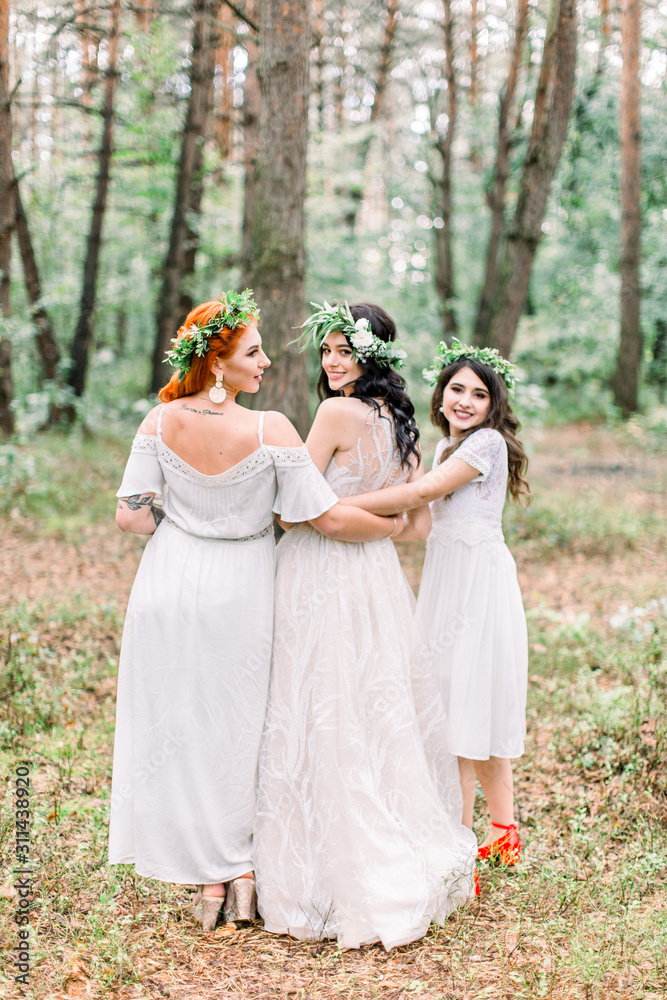 Back view of pretty cheerful bride and bridesmaids in white dresses and floal wreaths having fun at wedding day, walking in the pine forest.