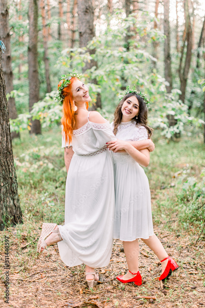 Funny two beautiful girls bridesmaids dance in the forest. Two bridesmaids in white dresses and wreaths on the heads smiling and having fun together Funny two beautiful girls bridesmaids dance in the forest. Two bridesmaids in white dresses and wreaths on the heads smiling and having fun together