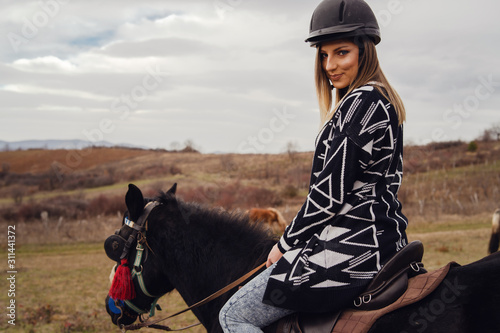 Young beautiful blonde caucasian woman female portrait against a gray cloudy sky in winter or autumn day wearing protective black helmet and sweater riding the horse back view © Miljan Živković