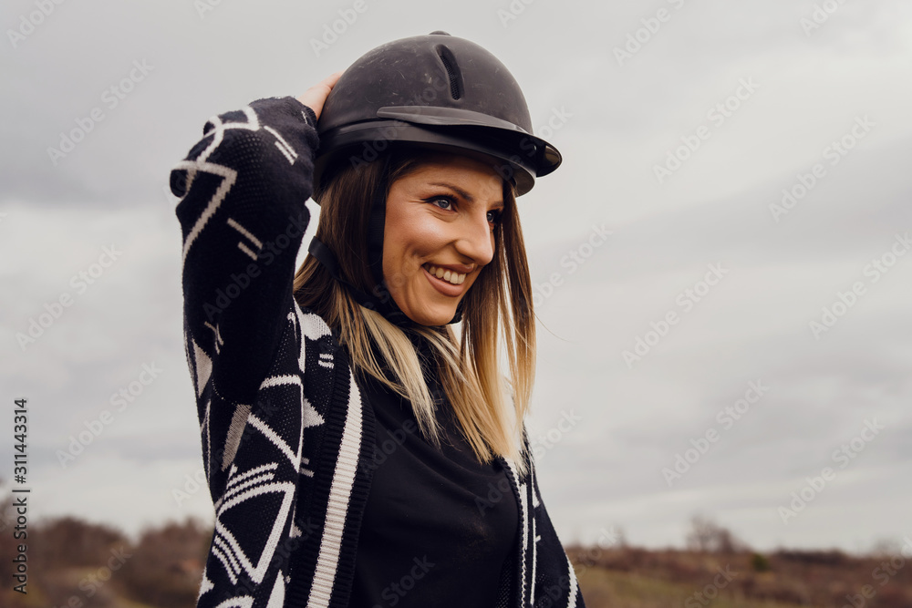 Young beautiful blonde caucasian woman female adjusting black protective helmet against a cloudy gray sky in winter or autumn day wearing sweater smiling