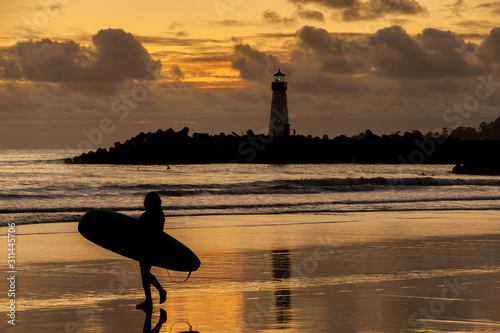Woman Surfer Walking Along the Beach in Front of a Lighthouse. A woman calls it quits after a long day of surfing during a beautiful sunset. Walton Lighthouse in the background. Santa Cruz, California
