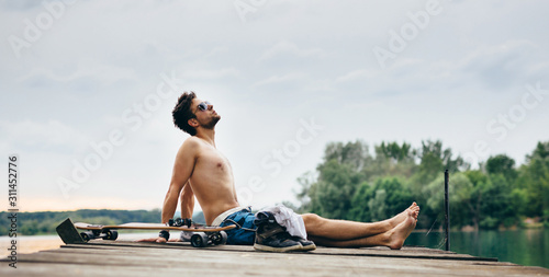 Young athletic man, in swimsuit, is sitting on wooden pier and sunbathing. Visible longboard, headphones and his clothes/sneakers are showing his style. Background and foreground out of focus.