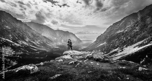 Wide landscape shot with mountains, valley and forest in background. Young couple standing on a rocky cliff in the middle and enjoying a magnificent view. Black and white shot with high dynamic range.