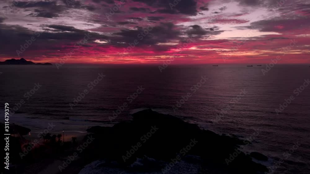 Aerial ascend over Arpoador rock dark in the foreground with the cloudy sky coloured red and magenta from the sun still beneath the horizon before sunrise Aerial ascend over Arpoador rock dark in the foreground with the cloudy sky coloured red and magenta from the sun still beneath the horizon before sunrise