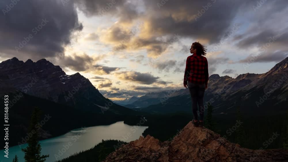 Time Lapse of Adventurous girl standing on the edge of a cliff overlooking the beautiful Canadian Rockies and Peyto Lake during a vibrant summer sunset. Taken in Banff National Park, Alberta, Canada. Time Lapse of Adventurous girl standing on the edge of a cliff overlooking the beautiful Canadian Rockies and Peyto Lake during a vibrant summer sunset. Taken in Banff National Park, Alberta, Canada.