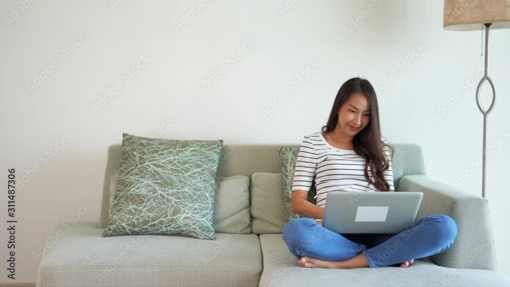 Young Asian woman sitting on stylish couch smiles as she types on her laptop. Copy space Young Asian woman sitting on stylish couch smiles as she types on her laptop. Copy space