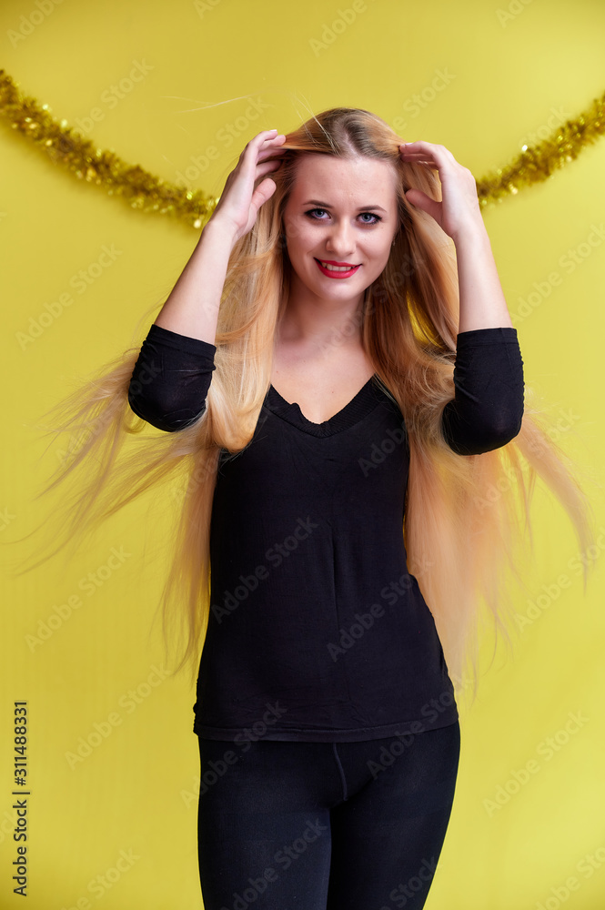 Naklejka premium Concept of a young blonde woman with New Year's decor. Portrait of a cute girl in a black T-shirt with long beautiful hair and great makeup. Smiling, showing emotions on a yellow background.