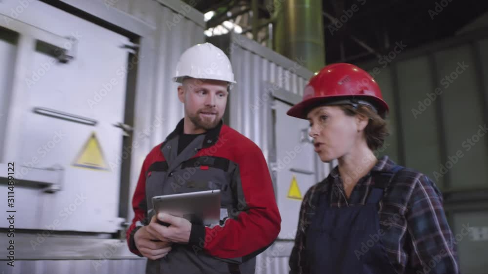 Chest-up shot of Caucasian man and woman in overalls and safety helmets walking together through production line machinery, engaged in conversation, listening to each other and talking Chest-up shot of Caucasian man and woman in overalls and safety helmets walking together through production line machinery, engaged in conversation, listening to each other and talking