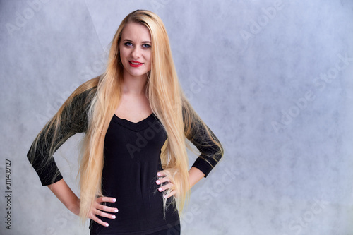 The concept of a blonde girl with a chic hairstyle. Portrait of a cute girl in a black T-shirt with long beautiful hair and great makeup. Smiling, showing emotions on a light gray background.