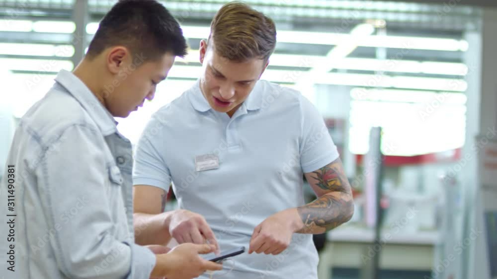 Medium shot of young tattooed male shop assistant helping Asian customer with smartphone at gadget store Medium shot of young tattooed male shop assistant helping Asian customer with smartphone at gadget store