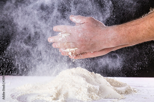Close up of chef human's hands preparing for kneading the dough on the table, powdering with flour. Making dough by hands at bakery or at home. Flour cloud in the air. Black background.