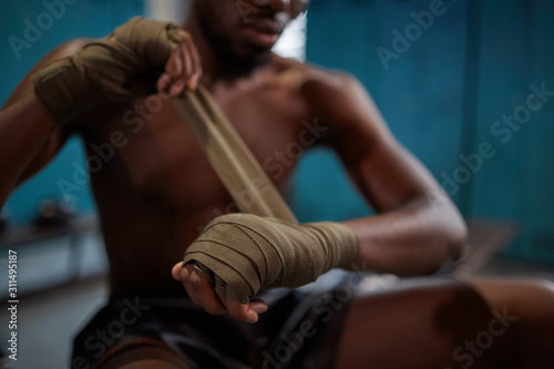 Close-up of African shirtless boxer sitting and wrapping bandage on his hands he is going to sports training