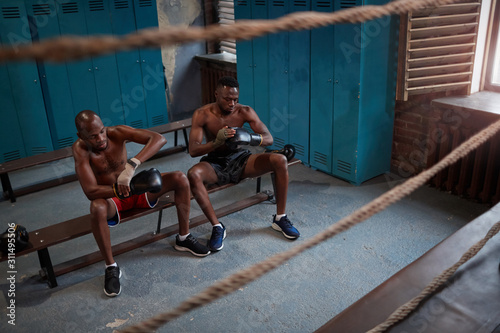 Two African boxers sitting on bench and wearing boxing gloves and preparing for sports training