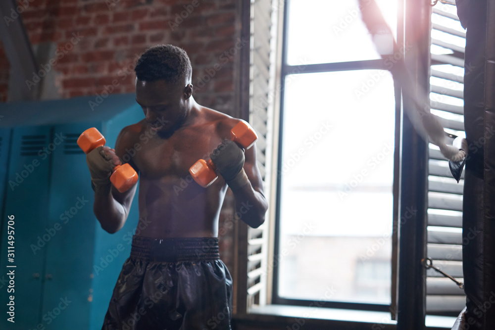 African young shirtless athlete standing and training with dumbbells in gym