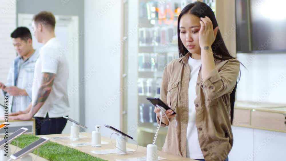 Portrait of young Asian woman in casual clothes choosing smartphones at gadget store and looking at camera, handheld Portrait of young Asian woman in casual clothes choosing smartphones at gadget store and looking at camera, handheld