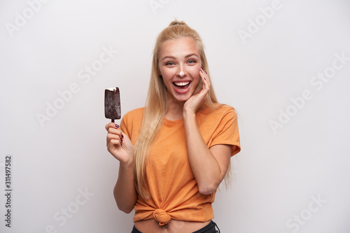 Indoor photo of happy attractive young long haired blonde woman keeping palm on her cheek and laughing happily while posing over white background, holding ice-cream in raised hand