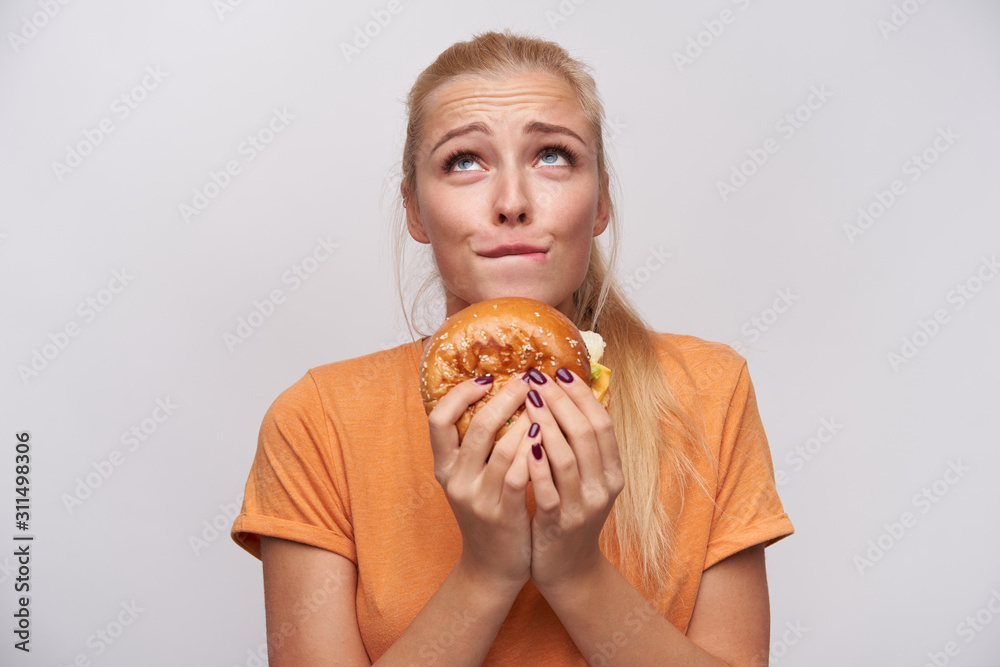 Young attractive long haired blonde woman with ponytail hairstyle wrinkling forehead and biting underlip while looking excusingly upwards with junk food in hands, isolated over white background