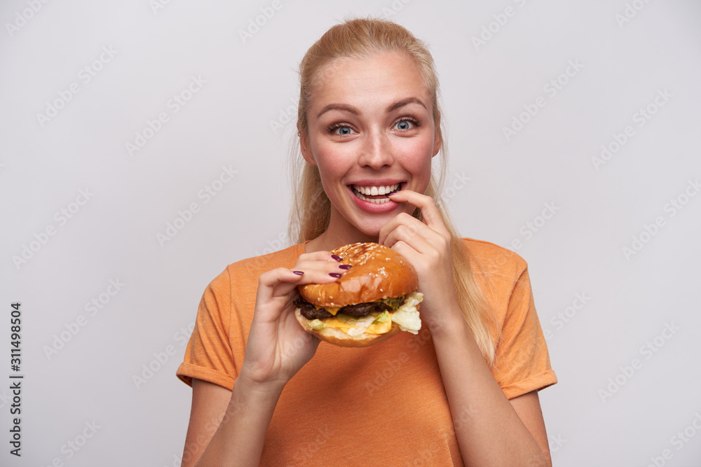 Cheerful blue-eyed young lovely blonde lady with casual hairstyle looking excitedly at camera and holding fresh hamburger in raised hand, keeping forefinger on underlip and smiling widely