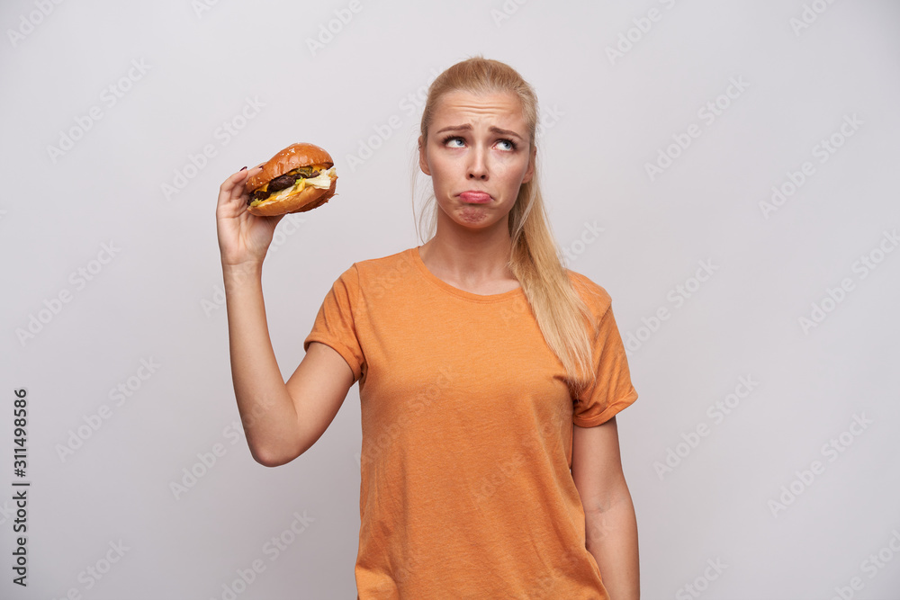Upset young long haired blonde lady in orange t-shirt looking aside sadly and wrinkling forehead while holding unhealthy food in raised hand, standing against white background Upset young long haired blonde lady in orange t-shirt looking aside sadly and wrinkling forehead while holding unhealthy food in raised hand, standing against white background
